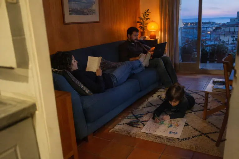 Dans un appartement marseillais des années 70, une femme lit allongée sur un canapé 3 places bleu marine, pieds sur son compagnon, leur fils dessine au sol