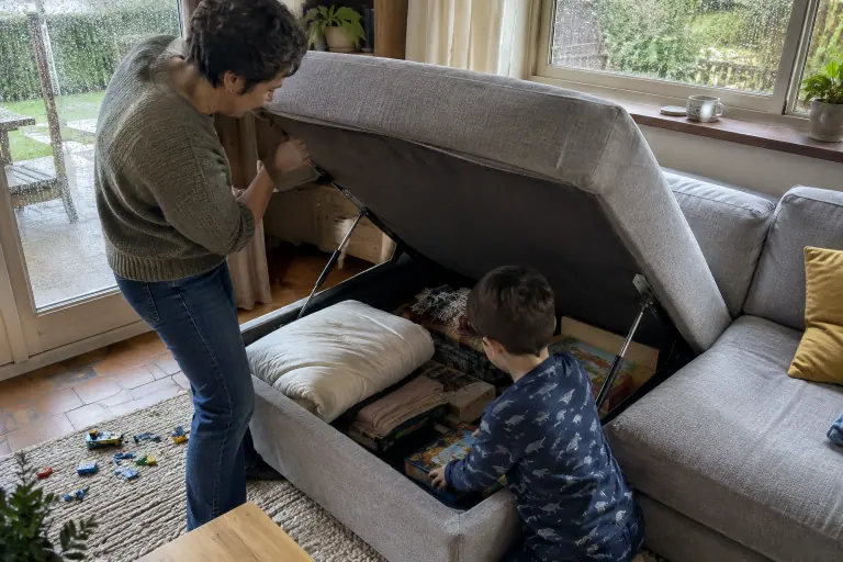 Une mère soulève l’assise de la méridienne d’un canapé d’angle gris clair pendant que son fils range un jeu de société dans le coffre