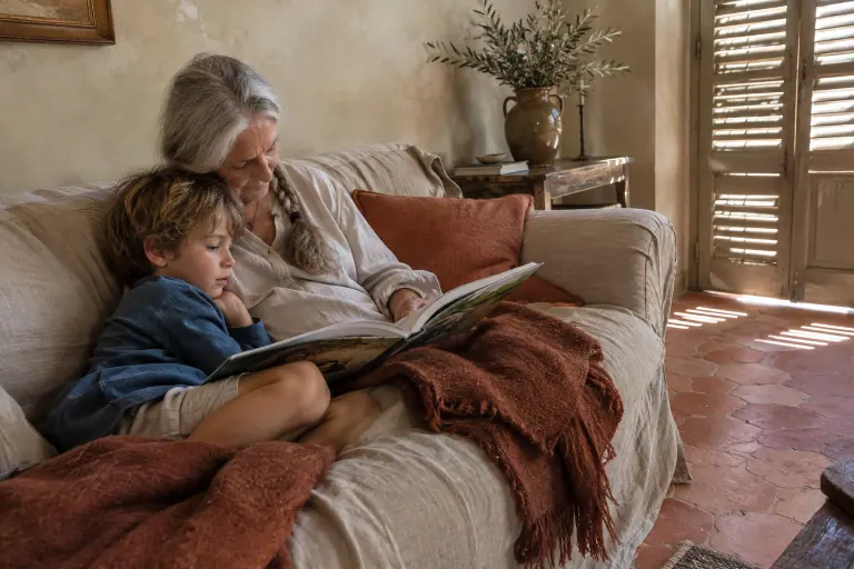 Une grand-mère lit un album à son petit-fils sur un canapé beige en lin, dans un mas provençal aux tomettes et aux murs à la chaux