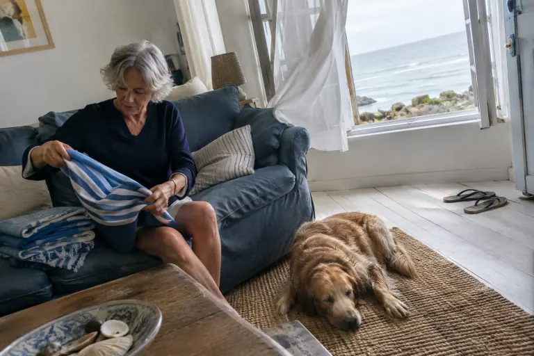 Une femme d’une soixantaine d’années plie des serviettes de plage sur un canapé en lin bleu jean, dans une maison de bord de mer normande