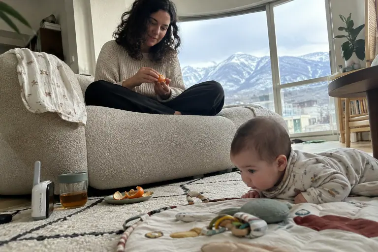 Une jeune mère épluche une clémentine assise sur un canapé arrondi en bouclette beige, son bébé allongé sur un tapis d’éveil, montagnes enneigées au loin