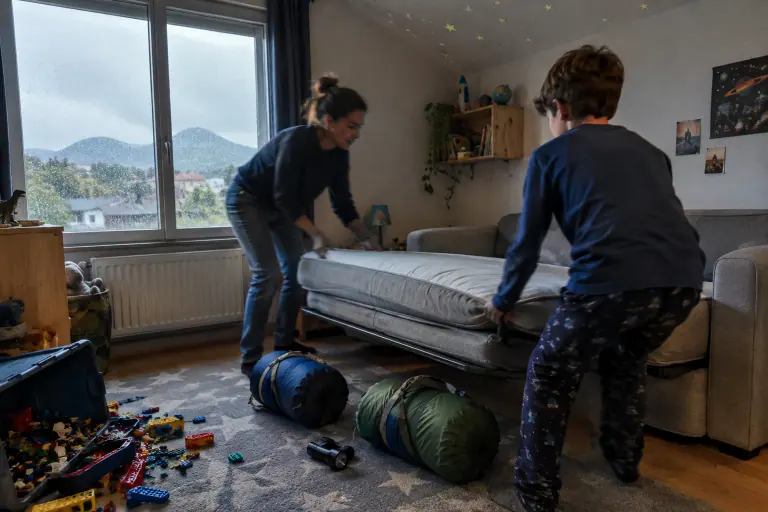 Une mère et son fils déplient en accordéon le matelas d’un BZ gris clair dans une chambre d’enfant, deux sacs de couchage prêts au sol