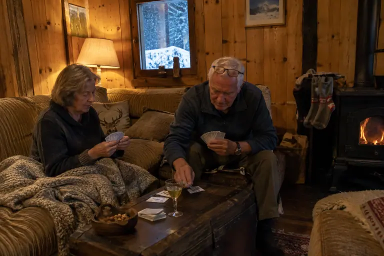 Dans un chalet savoyard, un couple de septuagénaires joue aux cartes devant un canapé en chenille couleur avoine, la neige tombant derrière la fenêtre