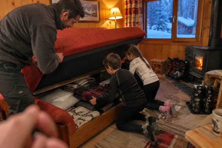 Un père soulève l’assise d’un clic-clac rouge brique pendant que ses deux enfants sortent une couverture du coffre, dans un appartement de Villeurbanne