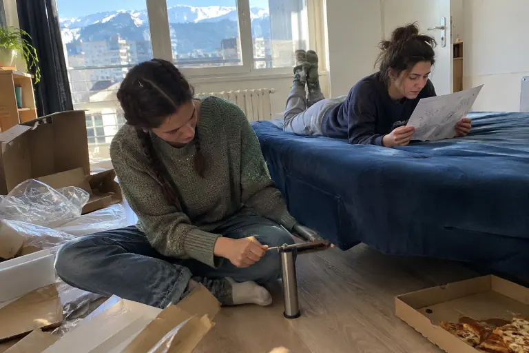 Une jeune femme visse les pieds d’un clic-clac bleu foncé au milieu des cartons, dans son premier appartement rennais, pendant qu’une amie lit la notice