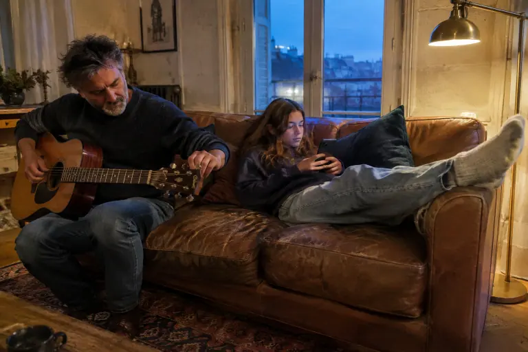 Un père accorde sa guitare sur un canapé en cuir cognac patiné pendant que sa fille ado regarde son téléphone, dans une maison bordelaise en pierre