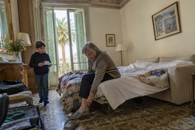 Une grand-mère enfile ses chaussons au bord d’un canapé convertible 3 places déplié dans un appartement dijonnais, son petit-fils lui apporte un café