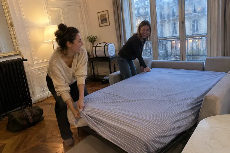 Deux amies installent un drap-housse rayé sur un canapé convertible gris clair tout juste déplié, dans un salon haussmannien un soir de pluie