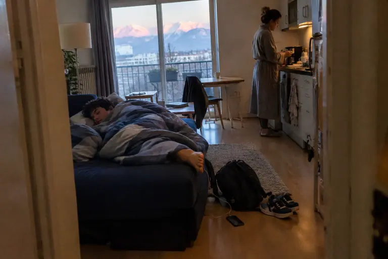 Un adolescent dort encore sur un canapé convertible bleu marine déplié dans le salon pendant que sa mère prépare le café, un matin d’hiver à Grenoble
