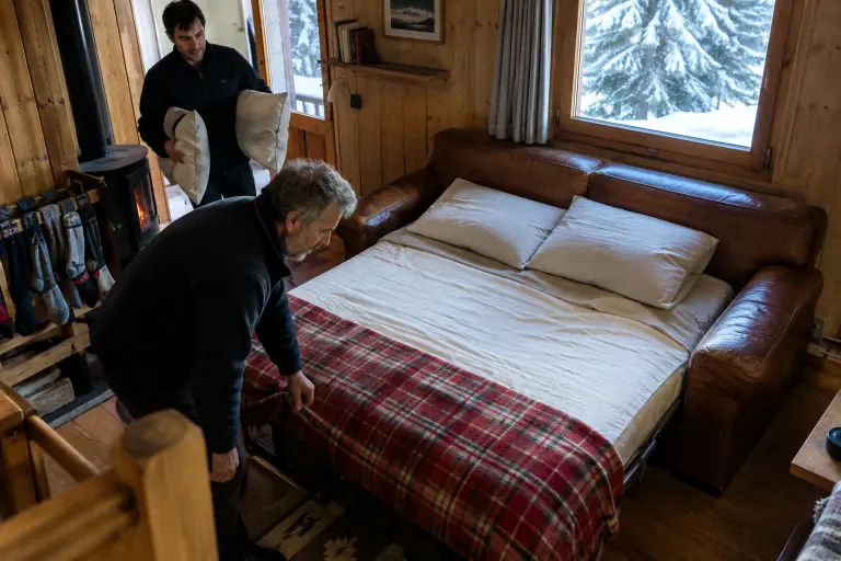 Un homme d’une soixantaine d’années borde une couverture sur un canapé-lit en cuir cognac déplié, dans un chalet savoyard en hiver