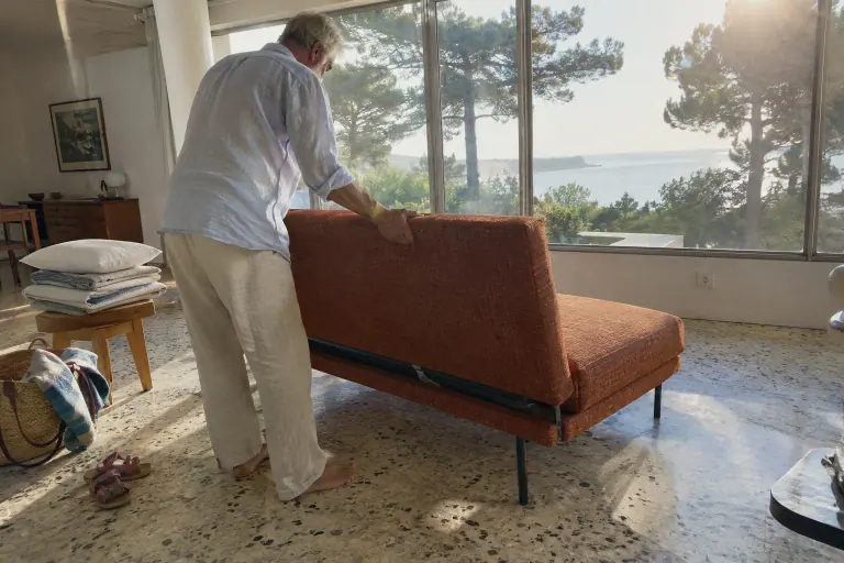 Dans une villa des années 50 à Royan, un homme relève le dossier d’un canapé convertible design orange brûlé à pieds métal noirs, posé au milieu de la pièce