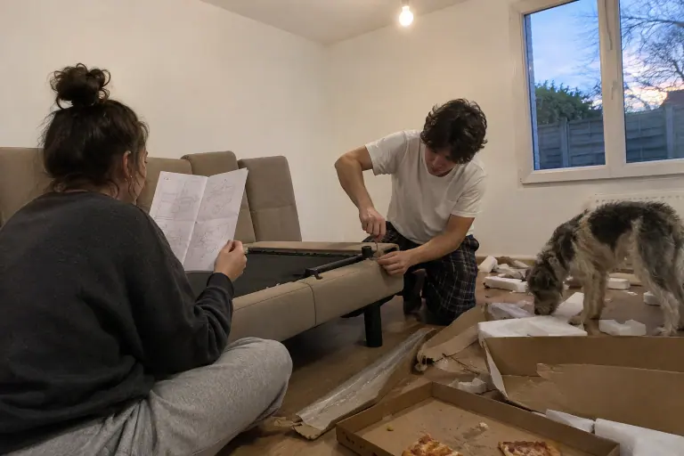 Un jeune couple monte un clic-clac taupe au milieu des cartons, dans un pavillon de banlieue, sous le regard curieux de leur chien