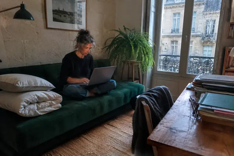 Une femme travaille sur son ordinateur, assise sur un clic-clac en velours vert émeraude, une couette pliée à côté, dans un bureau bordelais