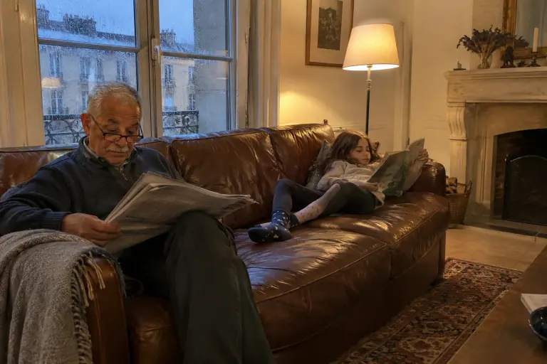 Un homme âgé lit le journal sur un canapé en cuir marron patiné, sa petite-fille allongée avec un livre à l’autre bout, un soir d’hiver
