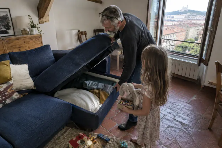 Un père soulève la méridienne d’un canapé d’angle bleu marine pour ranger un jeu dans le coffre, sa fille lui tend une peluche, sous un plafond à poutres