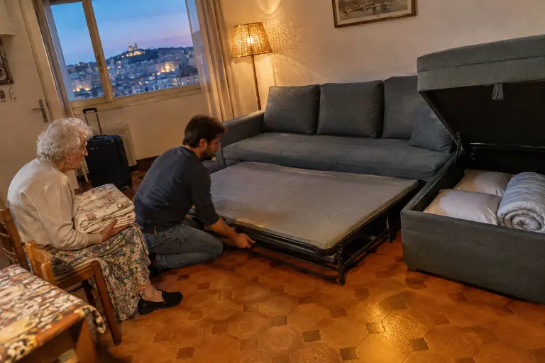 Dans un appartement marseillais des années 70, un homme déplie le couchage d’un canapé d’angle gris bleu tandis que sa grand-mère tient les draps