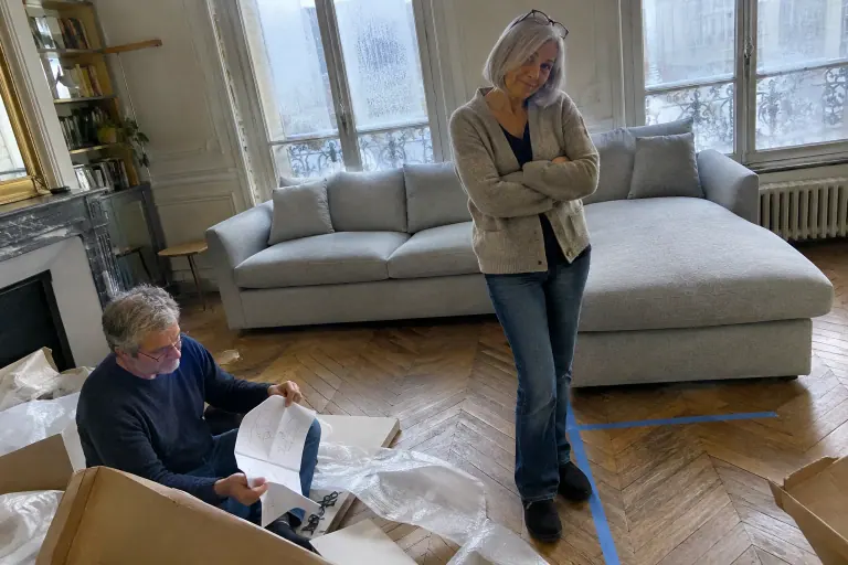 Une femme perplexe observe un canapé d’angle gris clair tout juste livré, méridienne à gauche, à côté d’un tracé au ruban adhésif prévu de l’autre côté