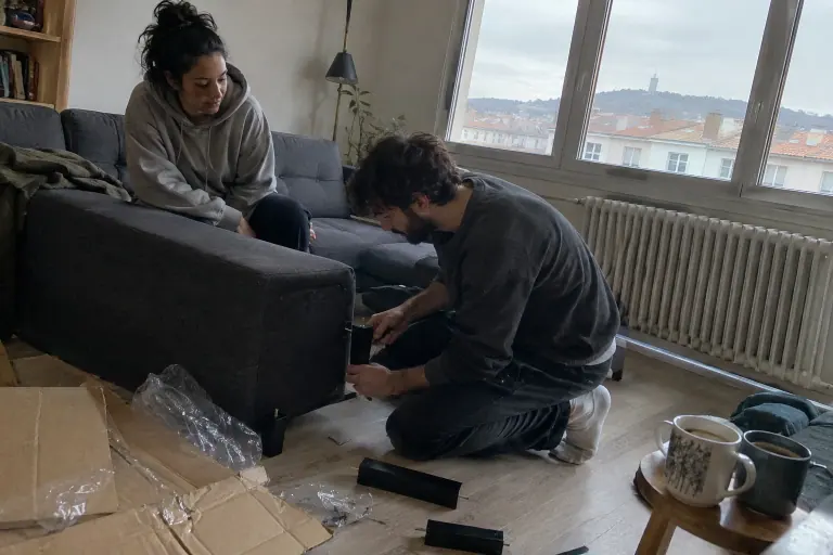Un jeune couple visse les pieds d’un canapé d’angle gris anthracite tout juste déballé, entouré de cartons, dans un petit appartement un matin d’hiver