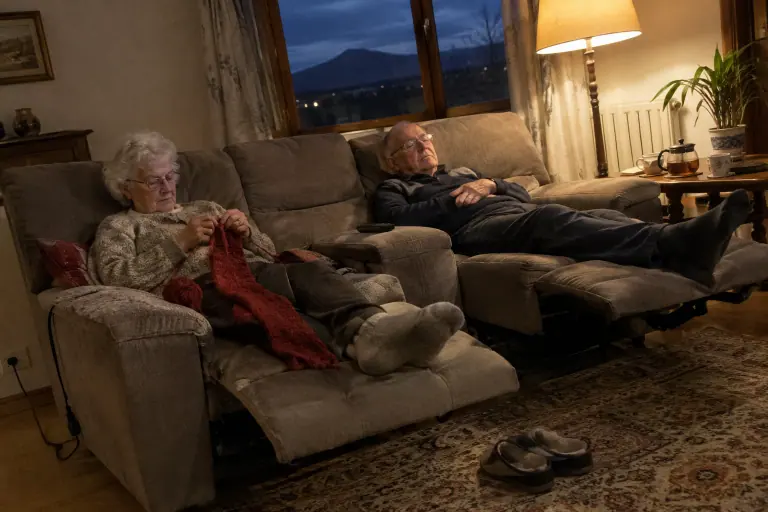 Un couple de retraités allongés sur un canapé d’angle relax taupe, repose-pieds levés, elle tricote pendant qu’il somnole devant un match