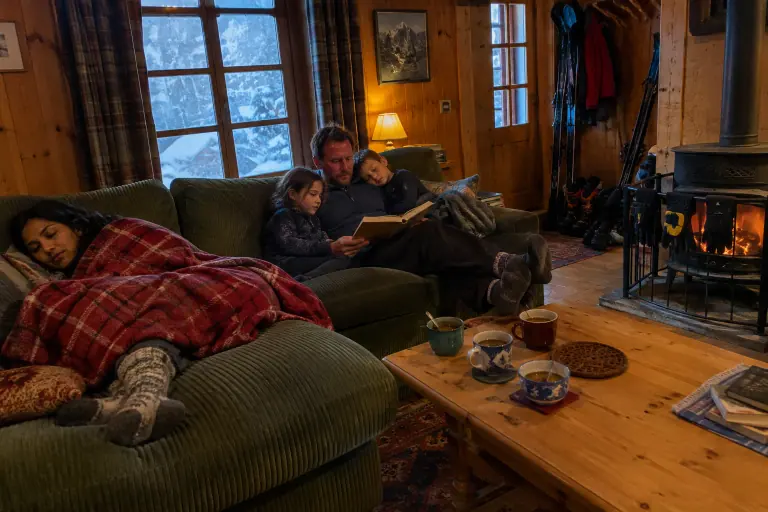 Dans un chalet savoyard sous la neige, une famille se repose après le ski sur un grand canapé d’angle vert sapin en velours côtelé, près du poêle