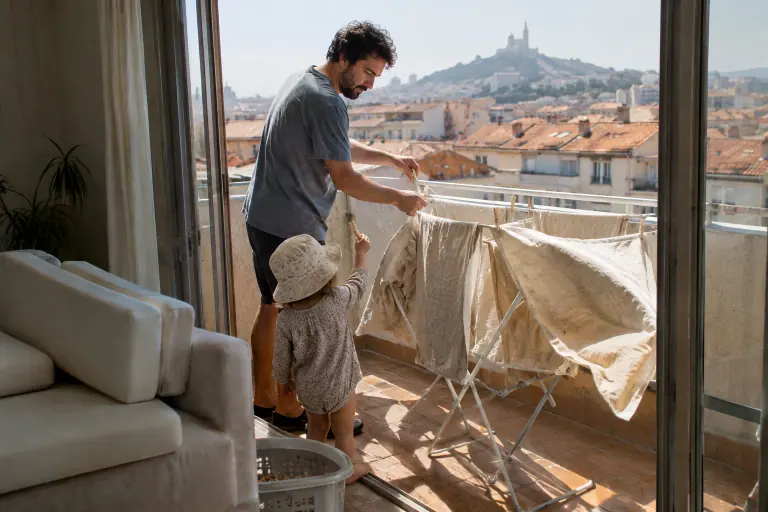 Sur un balcon marseillais, un père étend des housses de canapé couleur avoine tout juste lavées, aidé par son jeune enfant qui lui tend les pinces