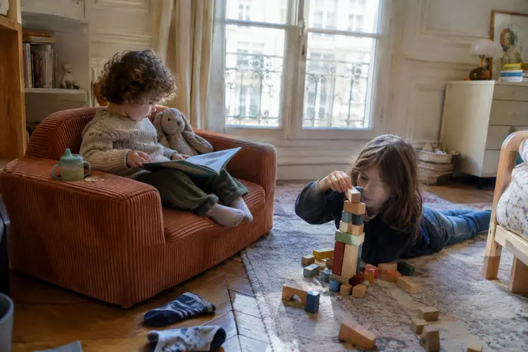 Un petit garçon de trois ans fait semblant de lire un album à son lapin en peluche, assis sur un mini canapé enfant en velours côtelé terracotta
