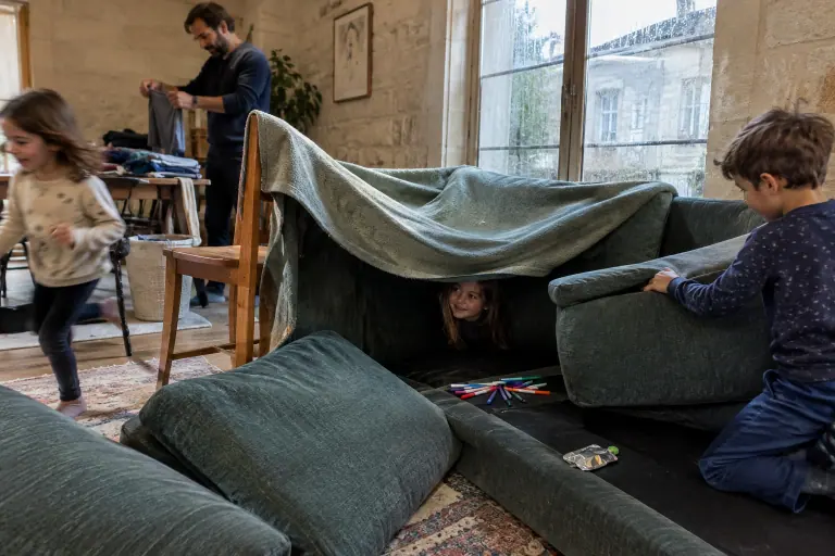 Trois enfants construisent une cabane avec les coussins d’un canapé d’angle vert chiné déhoussable, pendant que leur père plie du linge au fond