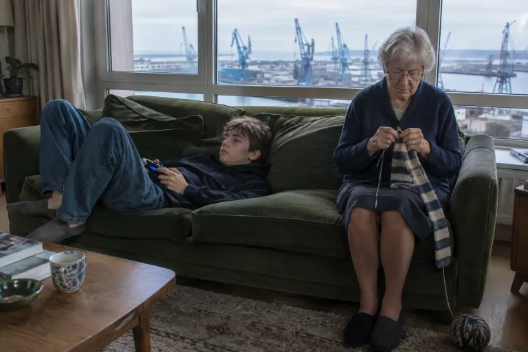 Un adolescent s’enfonce dans les coussins moelleux d’un canapé vert mousse tandis que sa grand-mère tricote, assise bien droite au bord de l’assise