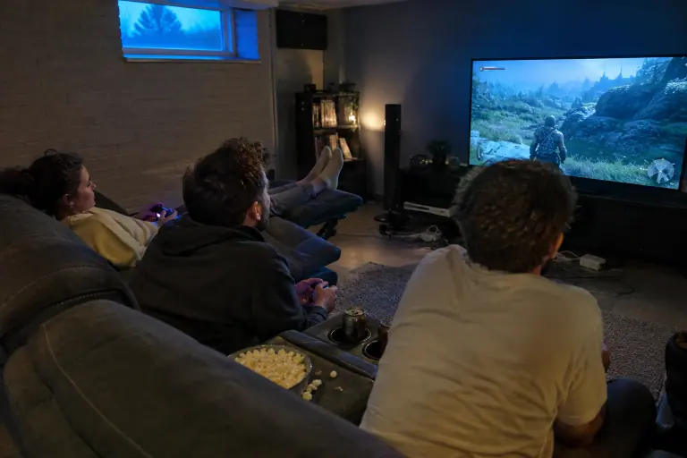 Trois amis jouent aux jeux vidéo sur un canapé home cinéma anthracite à console centrale, dans un sous-sol aménagé éclairé par la lueur de l’écran