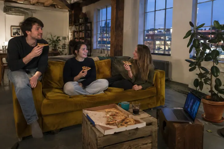 Trois colocataires mangent une pizza sur un canapé en velours jaune moutarde, dans un loft lyonnais aux grandes fenêtres un soir de semaine