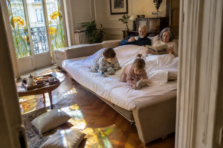 Des grands-parents prennent le café dans un large canapé lit beige déplié, leurs petits-enfants en pyjama à leurs pieds, dans un appartement de Nancy