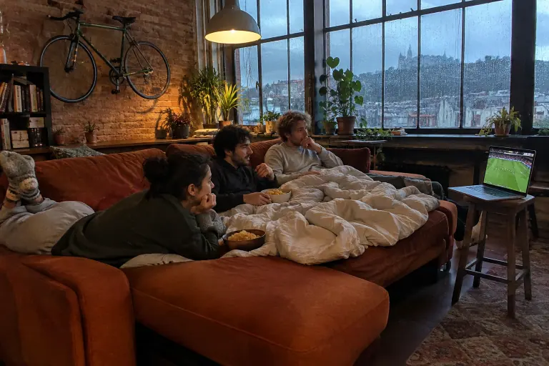 Trois colocataires regardent un match allongés sur un canapé rouille à méridienne ouvert en lit, dans un grand appartement de Nancy un soir de pluie