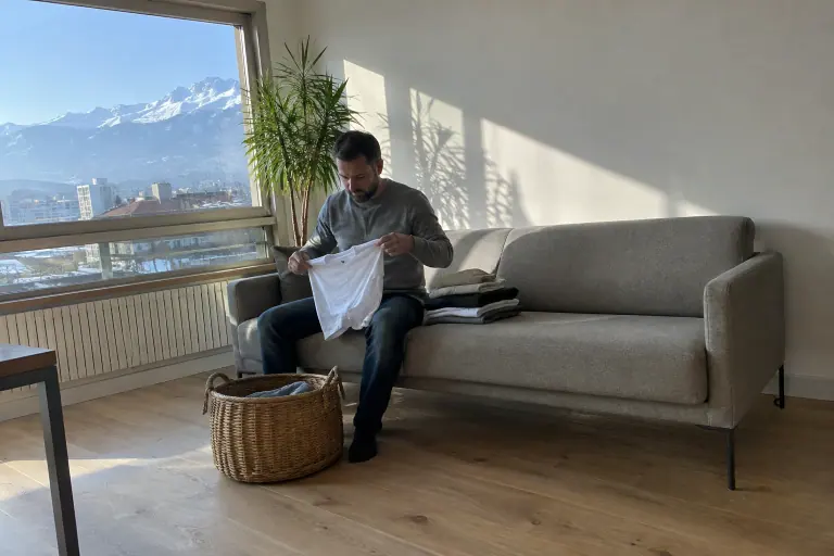 Un homme plie son linge au bord d’un canapé minimaliste greige à pieds métal fins, dans un salon presque vide avec vue sur des montagnes enneigées