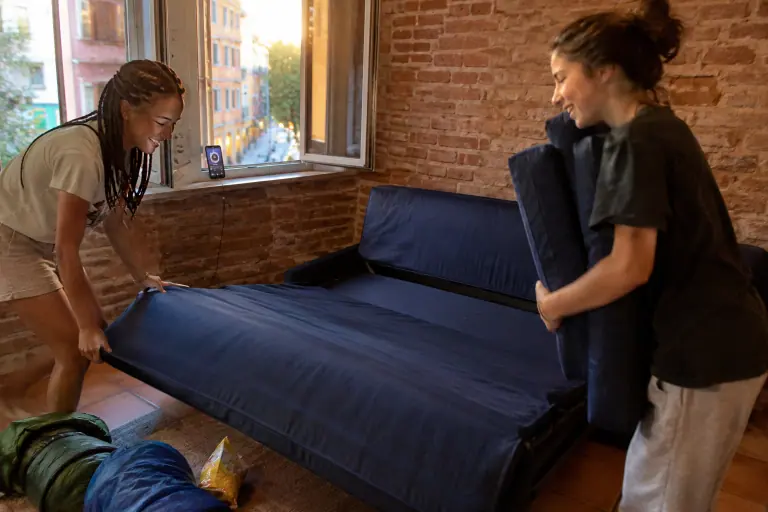 Deux adolescentes déplient en riant un canapé BZ bleu marine pour une soirée pyjama, dans une petite chambre en briques d’un appartement toulousain