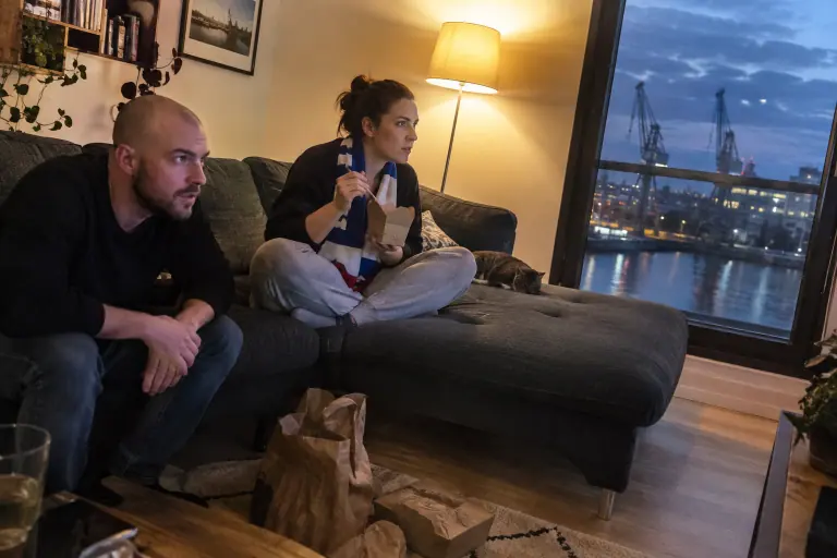 Un couple dîne de plats à emporter sur la méridienne d’un petit canapé d’angle gris foncé en regardant un match, un soir à Nantes