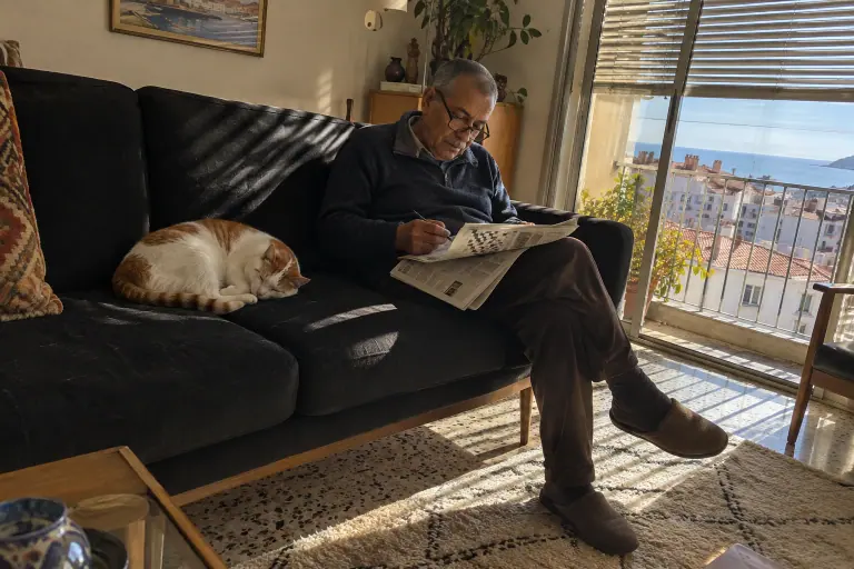 Un retraité fait les mots croisés sur un canapé noir à côté d’un chat blanc et roux endormi, dans un appartement marseillais des années 70