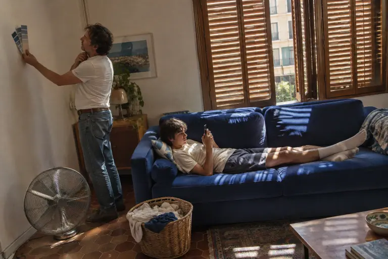 Dans un appartement marseillais des années 70, un homme compare des nuanciers de peinture derrière un canapé bleu cobalt où son fils est allongé