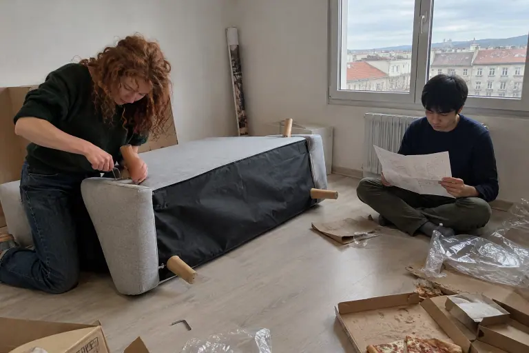 Un jeune couple monte un canapé en tissu gris clair renversé au milieu des cartons, dans un premier appartement, un samedi après-midi