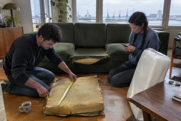Un jeune couple mesure la vieille mousse jaunie d’un coussin de canapé en velours côtelé vert affaissé, un bloc de mousse neuve posé à côté