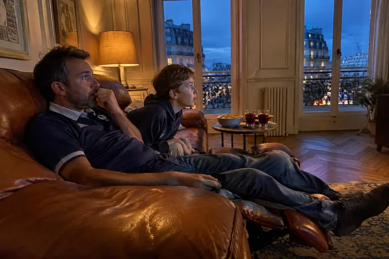 Un père et son fils regardent un match de rugby sur un canapé relax en cuir cognac au repose-pieds déplié, dans un appartement haussmannien