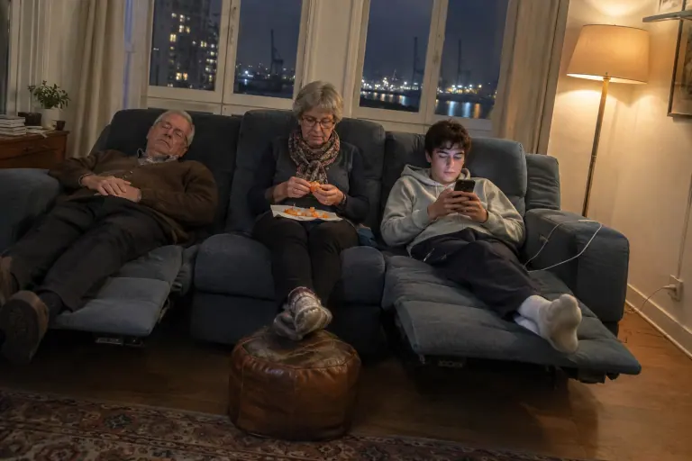 Un grand-père et son petit-fils allongés aux deux bouts d’un canapé relax électrique 3 places bleu ardoise, la grand-mère assise bien droite au milieu