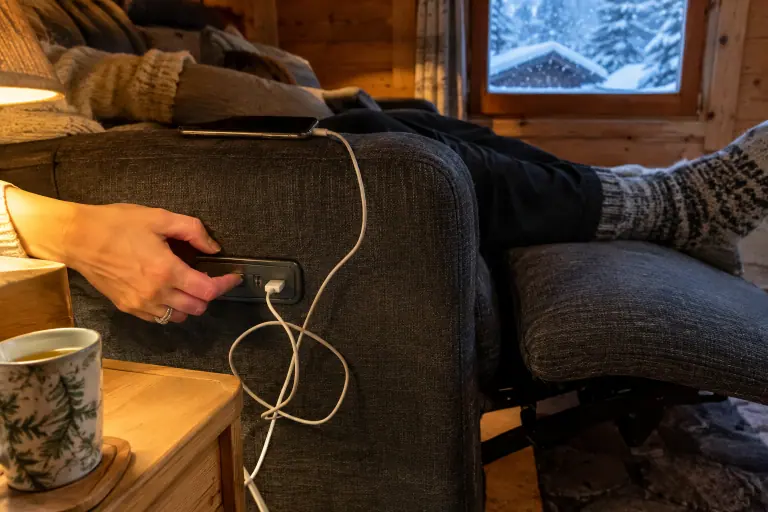 Gros plan sur une main qui appuie sur les boutons d’un canapé relax électrique gris foncé, un téléphone en charge sur le port USB de l’accoudoir