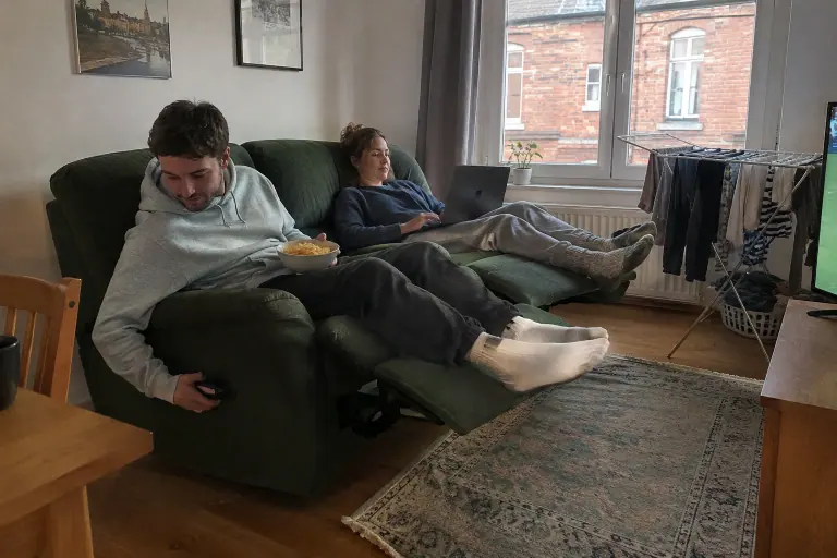 Un jeune homme tire la poignée latérale d’un canapé relax manuel vert foncé pendant que sa compagne est déjà allongée avec son ordinateur, dans un petit appartement