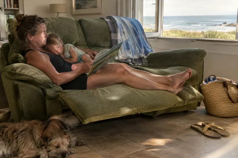 Une mère lit un album à son enfant blotti contre elle, repose-pieds déplié, sur un canapé relax en chenille vert mousse dans une maison en bord de mer