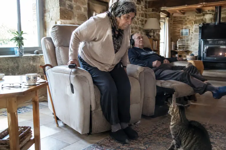 Une femme âgée se lève seule grâce au siège incliné d’un canapé releveur deux places beige, son mari se repose à côté, dans une longère bretonne