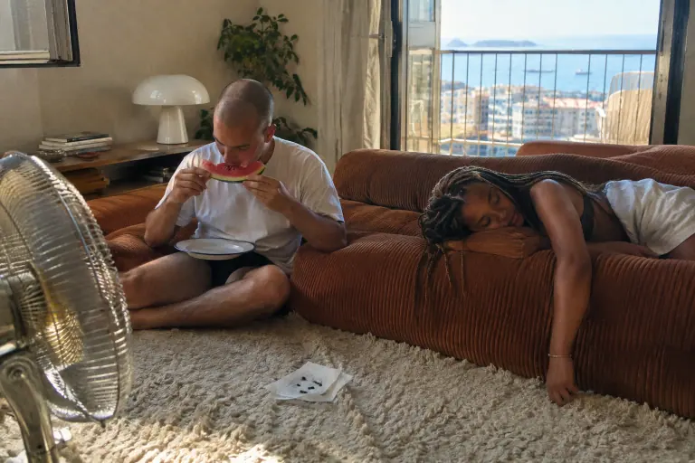 Deux colocataires sur un canapé bas en velours côtelé rouille : l’un mange de la pastèque, l’autre fait la sieste, dans un appartement marseillais
