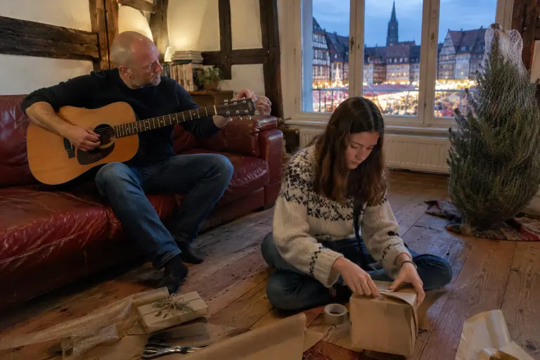 Un homme accorde une guitare sur un canapé en cuir rouge pendant que sa fille emballe des cadeaux, dans un appartement strasbourgeois à colombages