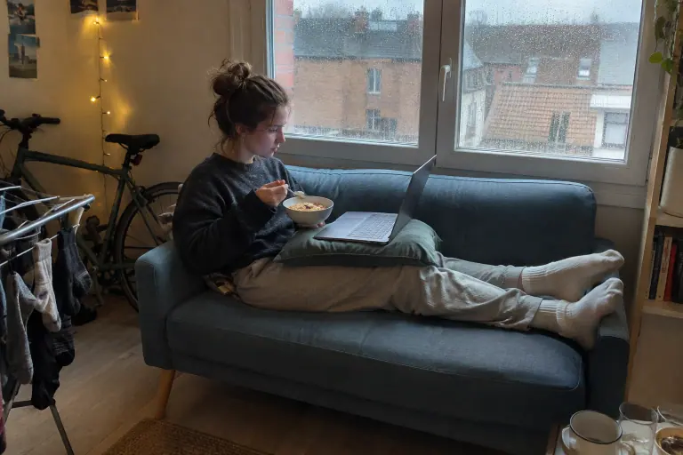 Une étudiante assise de travers, les jambes allongées sur un petit canapé sans accoudoirs bleu poudré, sous la fenêtre pluvieuse de son studio