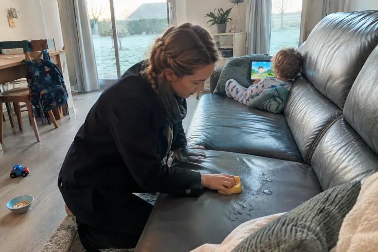 Une mère essuie du jus d’orange renversé sur un canapé en simili cuir gris foncé pendant que son enfant en pyjama regarde la télévision