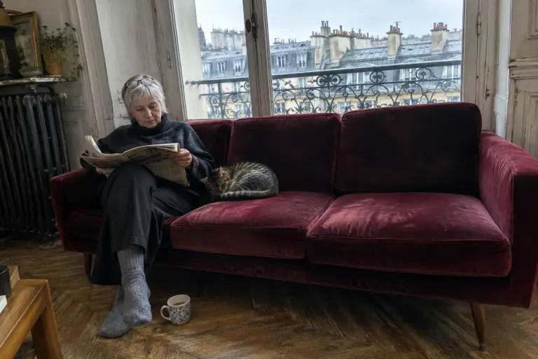 Une femme aux cheveux gris lit le journal sur un canapé en velours bordeaux, un chat tigré endormi à côté d’elle, dans un appartement haussmannien